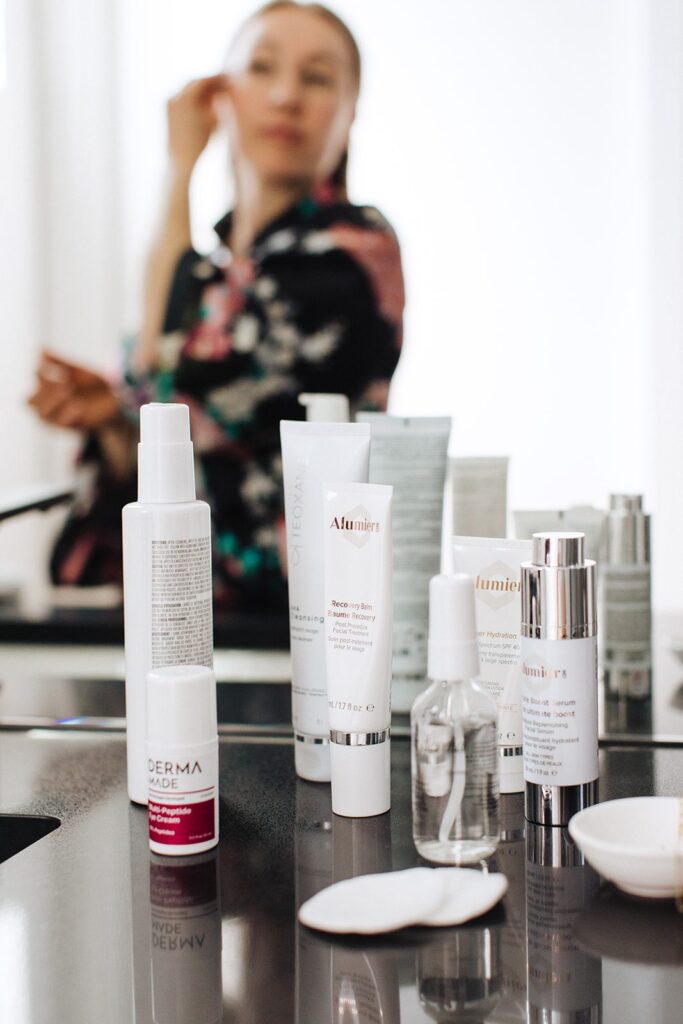 Skincare products sit on a counter as a woman applies a product to her face in the background.