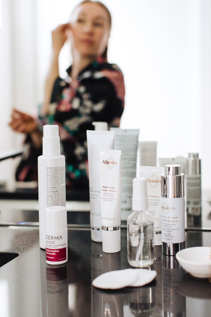 Skincare products sit on a counter as a woman applies a product to her face in the background.